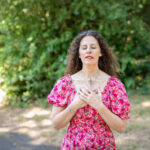 A woman with her hands on her heart using energy healing whilst standing amongst trees with a sunlit background